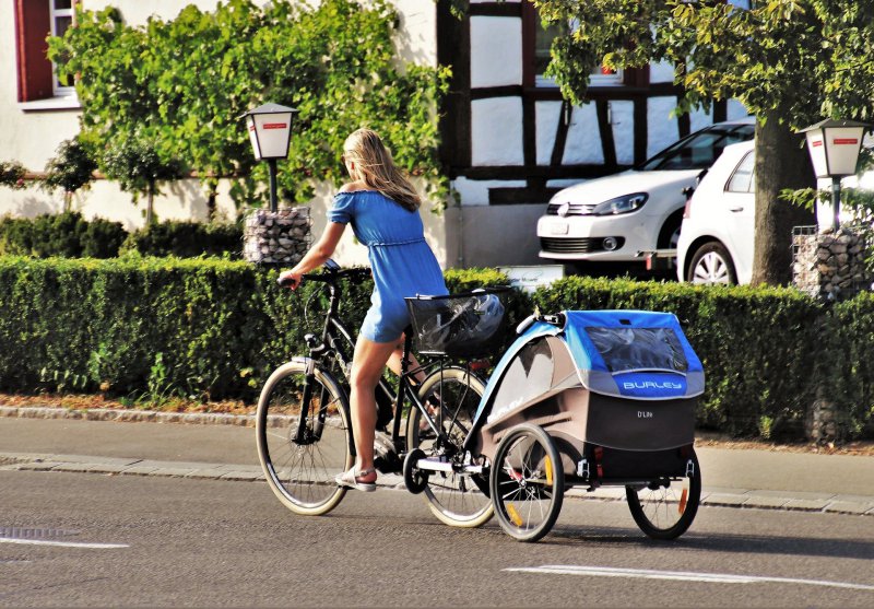 Eine Frau zieht ihren Fahrrad-Transportanhänger für Kinder mit ihrem Fahrrad hinter sich her.